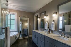 The Golden Locust Grey bathroom vanity with double sinks, marble countertop, large mirrors, and a freestanding tub by the window.