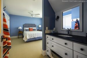 Heron Gate | Photography: © Daniel Vaughan (vaughangroup.ca) Blue bedroom with a patterned headboard and white bedding, looking toward a white bathroom vanity with dark tops.