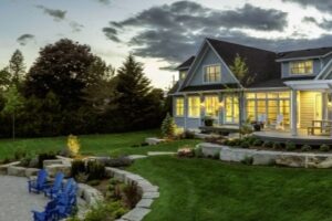 Heron Gate | Photography: © Daniel Vaughan (vaughangroup.ca) Panoramic view of a luxury grey shingle home at dusk, featuring a stone patio with blue chairs and a lake view.