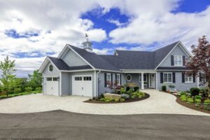 Heron Gate | Photography: © Daniel Vaughan (vaughangroup.ca) Grey shingle house with two white garage doors, a curved stone driveway, and rounded landscaped gardens under a blue sky.