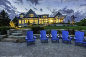 Heron Gate | Photography: © Daniel Vaughan (vaughangroup.ca) Rear view of a lit luxury home at dusk, featuring five bright blue Adirondack chairs on a stone patio by a lawn.