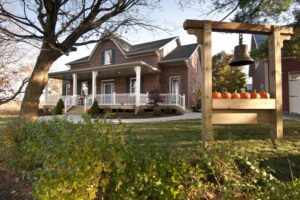 The Painted Maple A classic brick house with a wide front porch, seen behind a wooden post structure holding a large bronze bell.