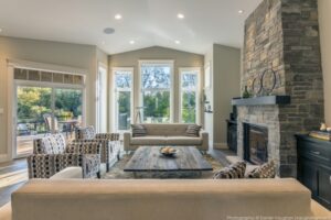 The Twisted Willow | Photography: © Daniel Vaughan (vaughangroup.ca) Light grey living room with a tall stone fireplace, patterned armchairs, and a large window view of trees.