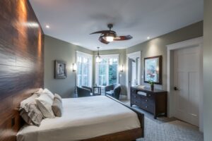 The Twisted Willow Modern bedroom featuring a dark wood accent wall, white bedding, a sitting area by the window, and a unique ceiling fan.