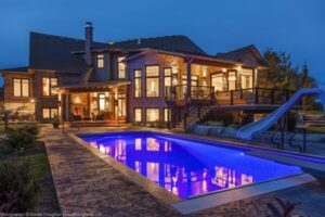 The Twisted Willow | Photography: © Daniel Vaughan (vaughangroup.ca) A large stone and brick luxury home at night, brilliantly lit, with a glowing blue pool and a water slide.
