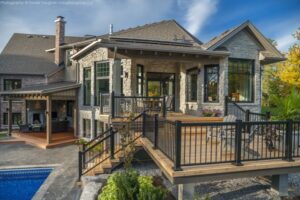 The Twisted Willow | Photography: © Daniel Vaughan (vaughangroup.ca) Stone and brick house featuring a multi-level wooden deck, black railings, and an outdoor patio by a blue pool.