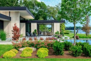 Modern flat-roofed patio with lounge chairs and sculptures, overlooking a manicured garden and a serene lake.