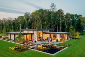 High-angle view of a modern wood-clad home at dusk, featuring glowing windows, reflecting pools, and a lush lawn.