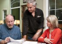 Man and woman sitting at table while looking at papers while another man stands in between them