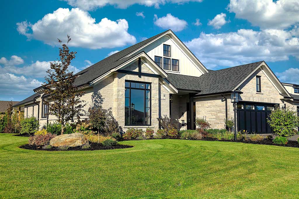 Contemporary stone and white-sided home with dark-framed windows, a black garage door, and a manicured lawn with garden beds.