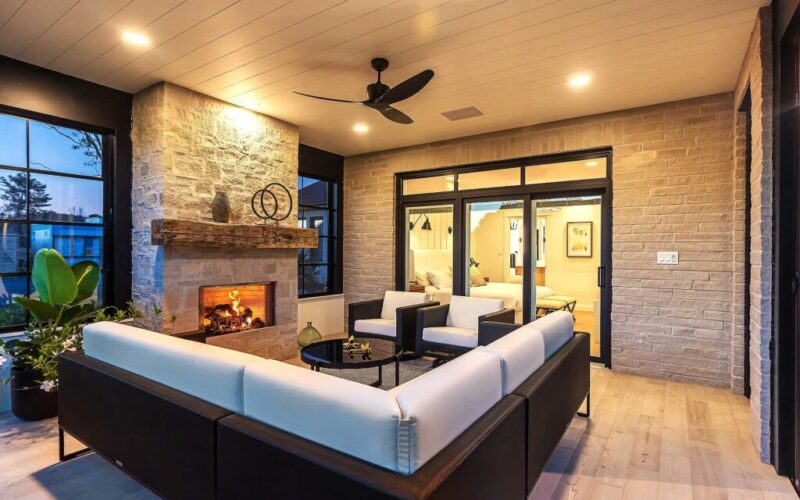 A cozy living room area with light wood flooring, a stacked-stone fireplace, a dark wicker sofa with white cushions, and large windows with black frames.