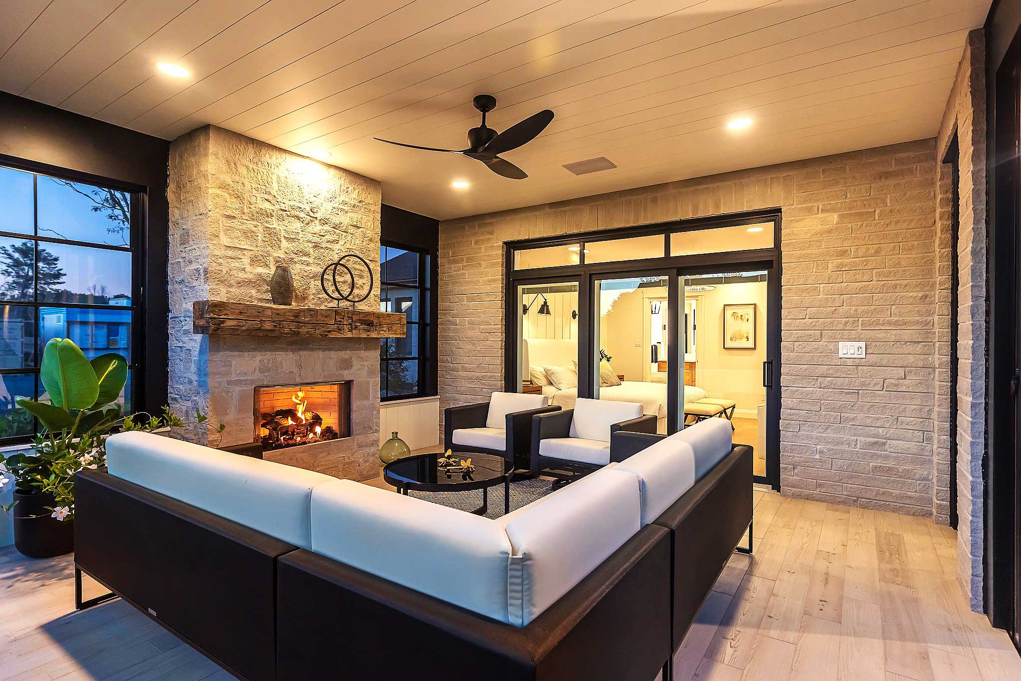 A cozy living room area with light wood flooring, a stacked-stone fireplace, a dark wicker sofa with white cushions, and large windows with black frames.