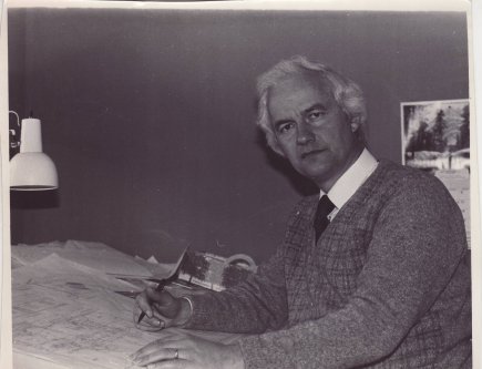 Black and white photograph of a man sitting at desk with large paper blueprint and a pen in hand