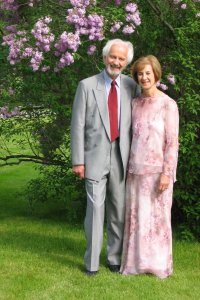 Senior couple dressed up in formal attire smiling for photograph with a blossomed purple flower tree behind them