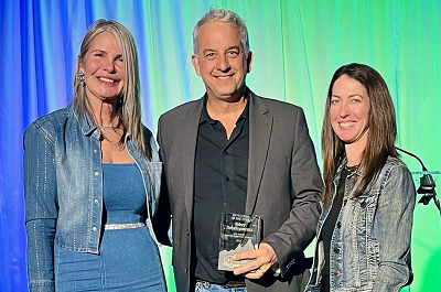 Three people smiling while a man holds a glass award in the middle of the two women with blue and green curtain background