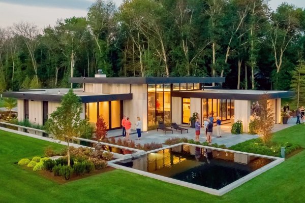 High-angle view of a modern wood-clad home at dusk, featuring glowing windows, reflecting pools, and a lush lawn.