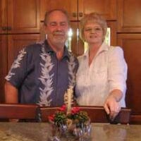 A couple standing for photo in kitchen with wood cupboards behind them and a granite tabletop with centrepiece in front of them