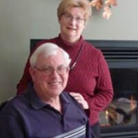 Wife stands behind sitting husband with her hand on his shoulder for a photo with a fireplace in the background