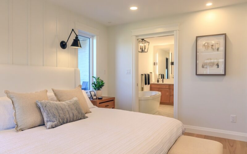 A clean white bedroom with decorative pillows on bed and light wood floors. A view of the master bathroom through can be seen through a doorway