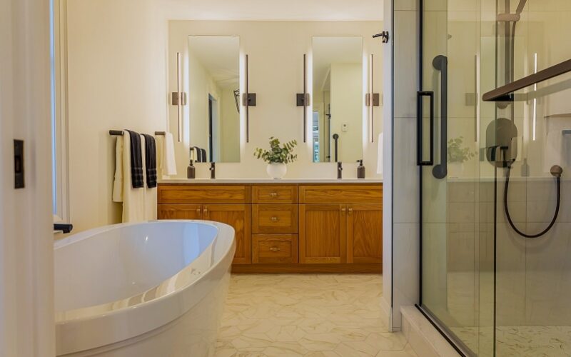 Modern Bathroom with standalone white tub and glass door shower. A two sink counter with long rectangular mirrors and long lights on each side of mirror are above each sink