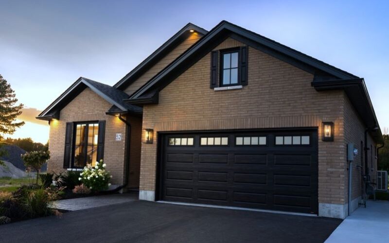 The front exterior of a tan brick bungalow at dusk, featuring a dark brown two-car garage, black window shutters, and glowing outdoor wall lanterns.