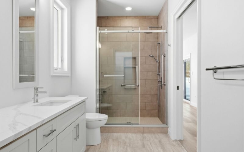 A clean, modern bathroom with a white marble-patterned vanity, a white undermount sink, and a walk-in shower with beige tiles and sliding glass doors.