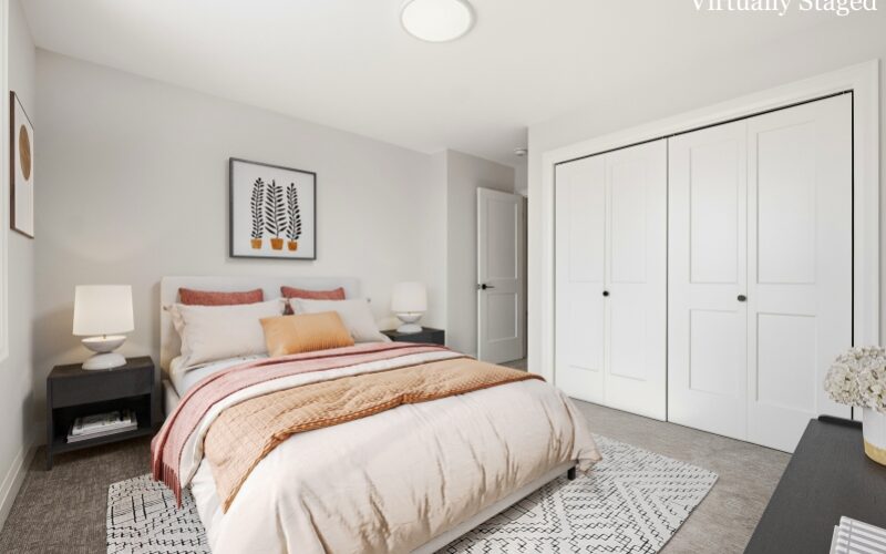 A cozy bedroom with white walls and grey carpeting, featuring a bed with coral and cream linens, dark nightstands with round white lamps, and a large white bi-fold closet.