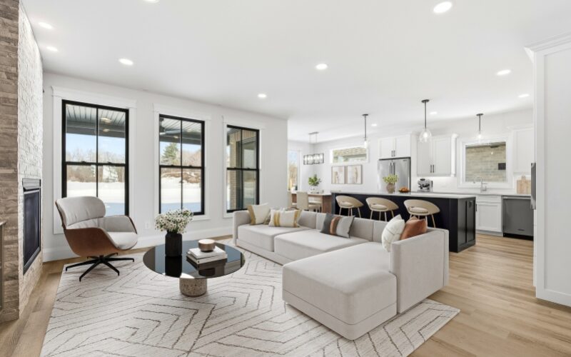 A wide shot of a contemporary living area and kitchen featuring a large L-shaped grey sofa, a black circular coffee table, and a kitchen island with black cabinetry and cream-coloured barstools.