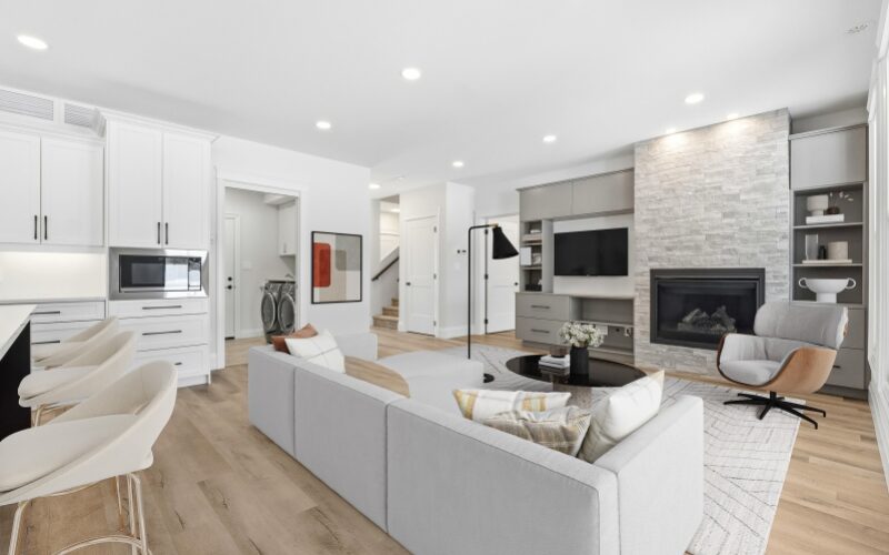 An open-concept living room and kitchen area showing a grey sectional sofa, a stone-faced fireplace with built-in shelving, and a glimpse of a laundry room in the background.