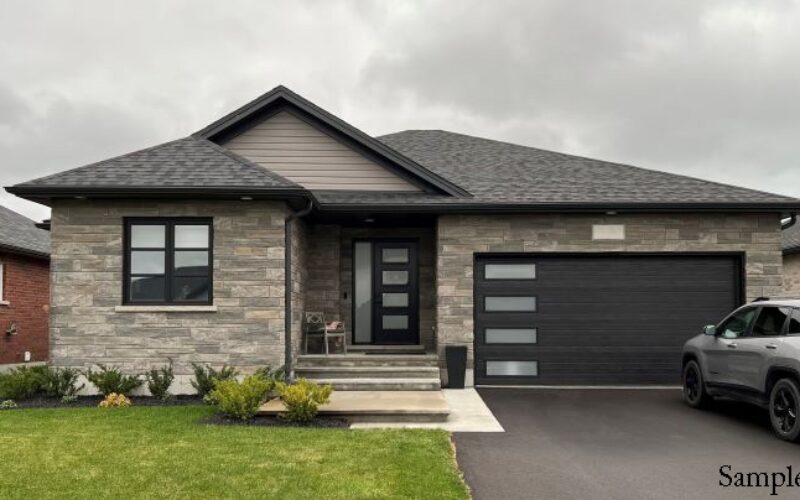 Modern single-story house with a stone exterior, black-framed windows, and an attached garage, featuring a small front porch with steps and a neatly maintained lawn under an overcast sky.