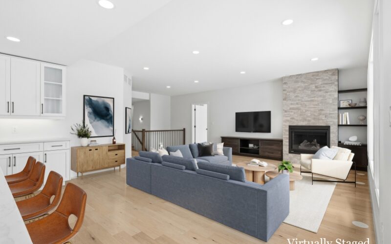 An open concept living room with a blue-grey L-shaped couch and fireplace with grey stone surround. White kitchen cabinets and brown stools are to the left.