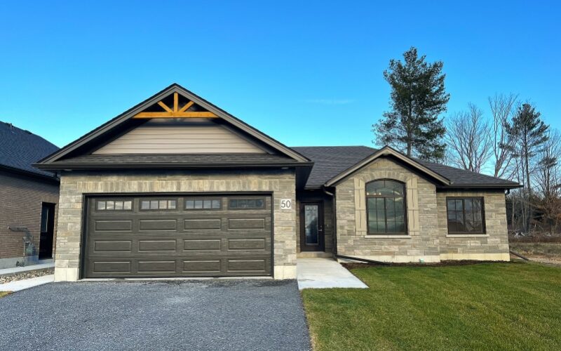 The front exterior of a modern grey stone bungalow with a dark grey two-car garage, a wooden gable accent, and black-trimmed windows under a clear blue sky.