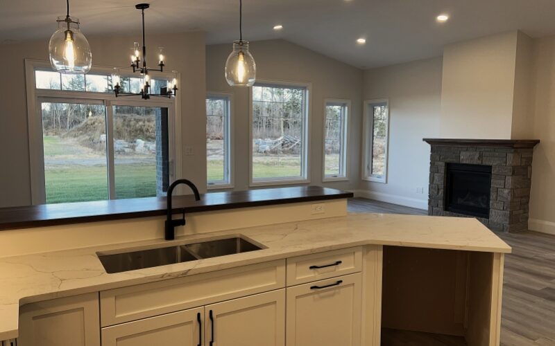A modern open-concept kitchen looking into a living area, featuring a white island with a marble-patterned countertop, double sink, and matte black faucet. The background shows a stone fireplace, large windows, and glass pendant lighting under a vaulted ceiling.