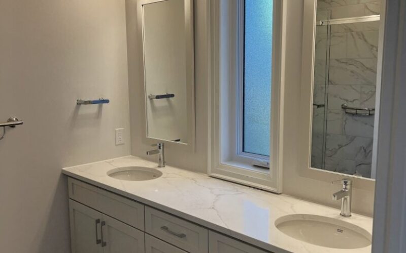 A modern bathroom featuring a long white double vanity with a marble-patterned countertop and two undermount sinks. Above each sink is a rectangular white-framed mirror topped with a three-light chrome fixture. A frosted vertical window sits between the two mirrors, and the room is finished with neutral grey walls and large slate-colored floor tiles.