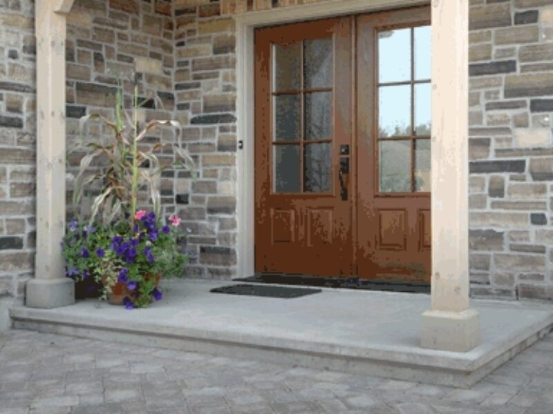 Front door entrance of stone house with interlock walkway up to a concrete step to two wooden doors with glass panels. The overhang is supported by two wooden posts and brackets