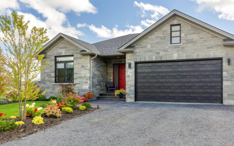Front view of a modern stone bungalow with a dark wood-style two-car garage, a vibrant red front door, and a manicured garden with colourful flowers.