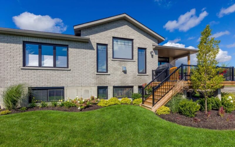 The back of a light grey brick house featuring a raised wooden deck with black railings, large dark-framed windows, and a lush green lawn.
