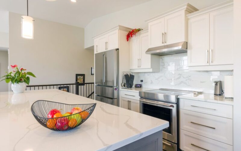 Close-up of a kitchen island with a marble-patterned countertop and fruit bowl, looking toward stainless steel appliances and white cabinetry with a marble backsplash.