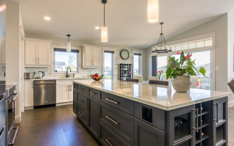A wide view of a modern kitchen featuring a large dark grey island with white quartz countertops, pendant lighting, and an open layout leading to a bright dining area.