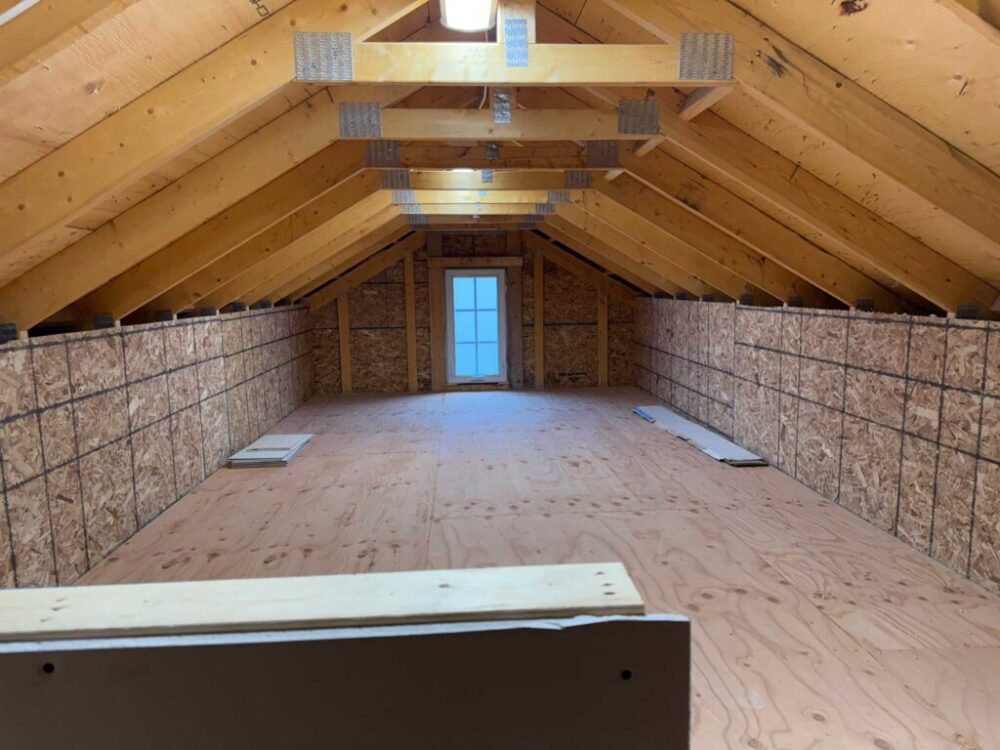 An unfinished attic or loft storage space with exposed wooden roof trusses, plywood flooring, and particle board knee walls. A small window at the far end provides natural light to the long, narrow space.