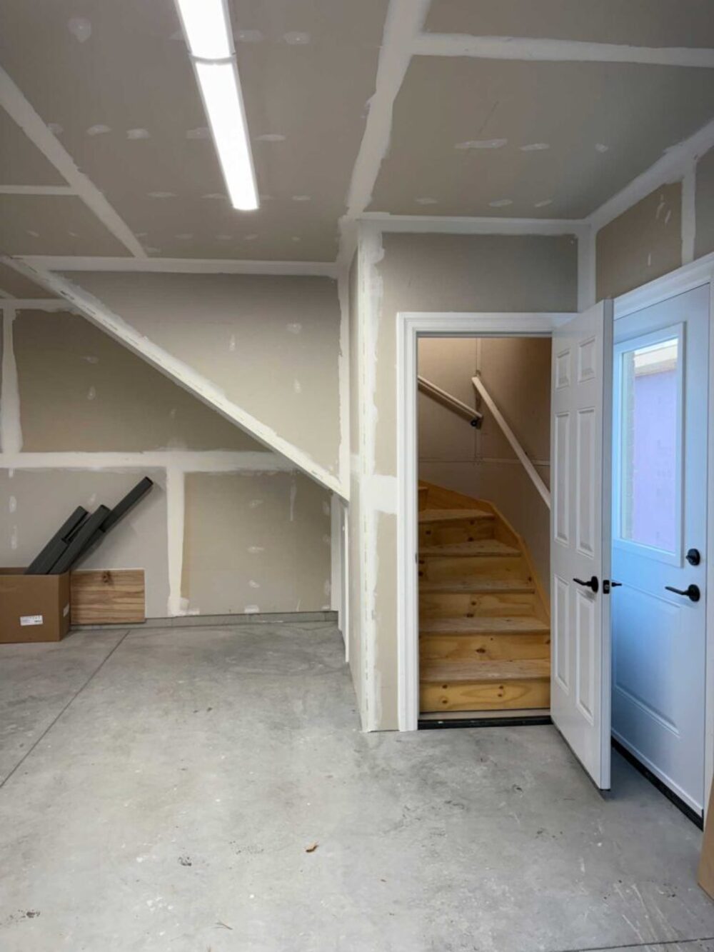 The interior of an unfinished garage with taped drywall and a concrete floor. An open white door reveals a set of unfinished wooden stairs leading upward, situated next to a second white exterior door.